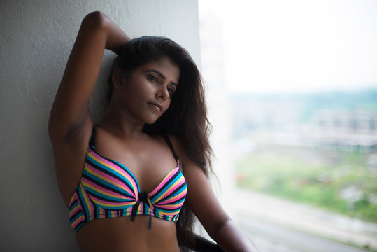 Portrait of an young and beautiful dark skinned Indian Bengali woman in colorful lingerie/bikini and hot pants posing in casual mood on a balcony in white urban background. Boudoir photography.