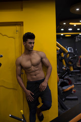 Naklejka premium Portrait of an young and handsome Indian Bengali brunette man with muscular body standing in a vibrant yellow background in a multi gym. Fitness and Indian lifestyle.