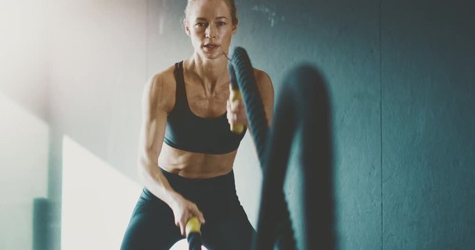 Fit attractive woman working out with battle ropes in the gym, intense focused woman training hard to achieve her fitness dreams