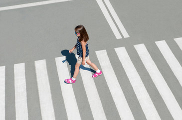 Stylish child in glasses, fashion clothes walking along summer city crosswalk. Kid on pedestrian side walk. Concept pedestrians passing a crosswalk. From top view. Behind. Shadow at zebra crossing