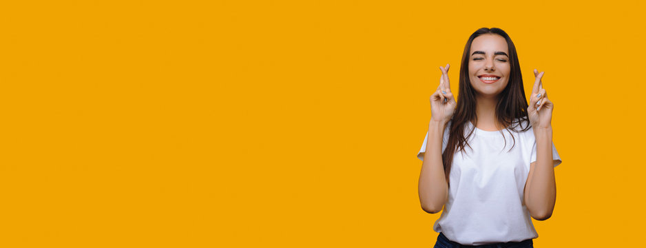 Cheerful caucasian woman posing aside with crossed fingers on yellow wall with blank space