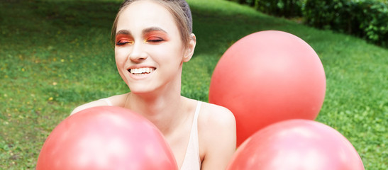 Pretty young woman with bright make up. Outdoor green portrait. Red balloons at park. Happy model face. Orange mascara artist style. Fashion smokey