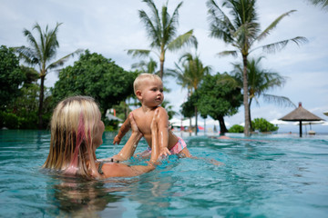 Mother and baby play in outdoor swimming pool of luxury spa resort in Bali. Summer vacation for family with children. Kids in hot tub outdoors with ocean view.