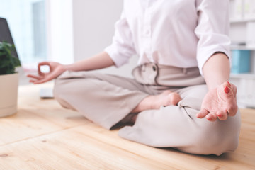 Obraz na płótnie Canvas Close-up of unrecognizable businesswoman sitting in lotus position and holding hands in mudra while practicing relaxing meditation at workplace