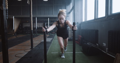 Motivation concept, young athletic blonde woman pushing heavy training sled towards camera training in gym slow motion.