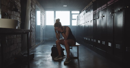 Camera follows athletic European woman entering dark gym locker room, sitting along feeling tired and upset slow motion.