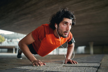 Confident handsome sporty young determined fit man listening music on earphone doing push ups at...