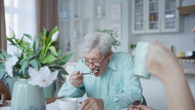 Panning shot of four cheerful elderly friends, two men and two women, enjoying joyful talk over tea and desserts sitting at dining table at home