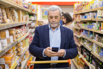 Man with shopping trolley using smartphone in store. Focused bearded mature man holding mobile phone and choosing goods in grocery store. Shopping and technology concept