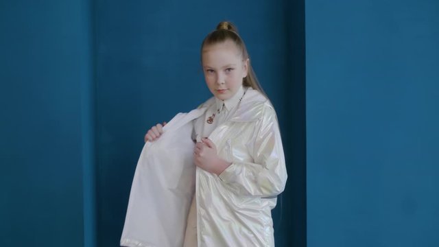 Teenager girl model posing in white jacket on blue wall background. Fashion teenager girl posing in stylish white coat on podium in studio