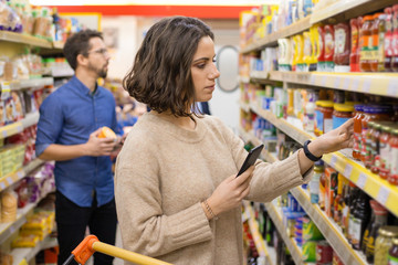 Woman holding smartphone and choosing goods in supermarket. Young woman reading checklist via smartphone and choosing products in grocery store. Supermarket concept