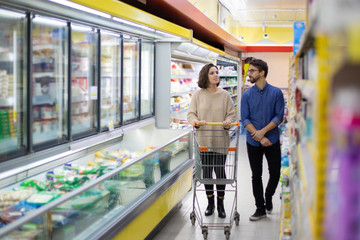 Couple walking and talking in grocery store. Happy young man and woman with shopping trolley choosing products in supermarket. Shopping concept