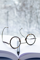 Round glasses and a black pencil lie on a notepad with white pages against the background of the landscape outside the window