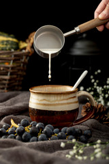 Man Pouring Milk In A Brown Coffee Cup With Grapes On A Grey Cloth