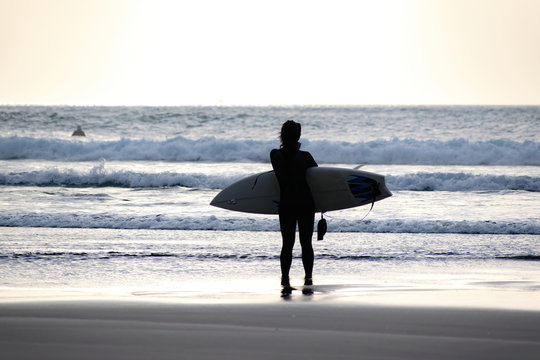 View Of Young Woman Surfer Wearing Black Wetsuit And Holding Surfboard, Preparing To Enter Piha Beach Surf