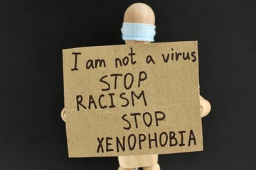Cardboard sign with text in the hands of a wooden doll or mannequin in a medical mask. Concept of anti racism, anti xenophobia, the social situation around the Coronavirus or Covid-19.