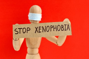 Cardboard sign with text stop xenophobia in the hands of a wooden doll or mannequin in a medical mask. Concept of anti racism, anti xenophobia, the social situation around the Coronavirus or Covid-19. - obrazy, fototapety, plakaty