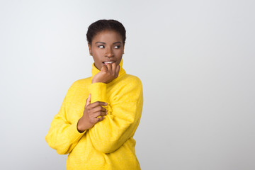 Curious young woman looking aside. Beautiful interested young African American woman in yellow knitted sweater standing with hand near face and looking away on white background. Curiosity concept