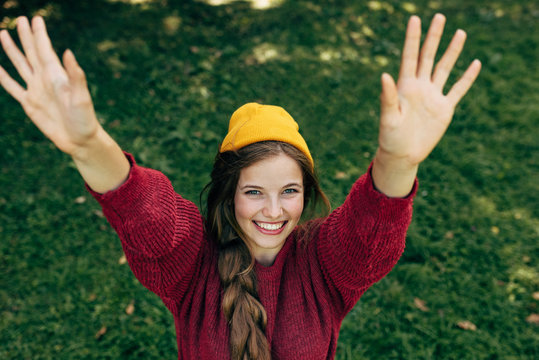 Top view image of a happy blonde young woman smiling broadly with a healthy toothy smile, with raised arms up, wearing a red sweater, and yellow hat, posing on green grass background in the park.