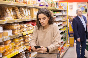 Woman using smartphone in grocery store. Young woman reading checklist via smartphone and choosing goods in grocery store. Supermarket concept