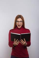 Excited female college student reading book. Cheerful young woman in eyeglasses holding notebook and looking at camera. Education concept