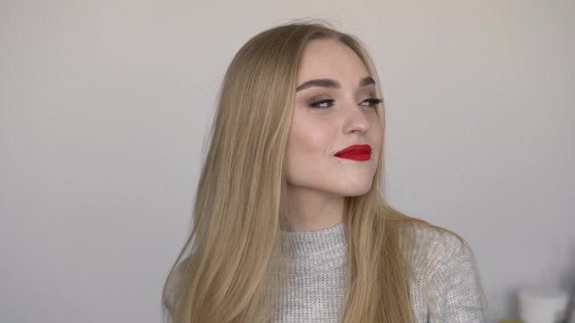 Indoors slow motion closeup face shot of young smiling woman smoothing her long blond hair by turning head, bright red lips, dressed grey sweater, posing against white wall natural light