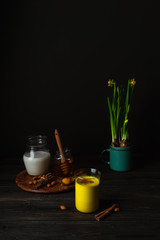 Fototapeta na wymiar Vegan turmeric latte in a glass, almond milk, spices, potted yellow duffodils on dark rustic background