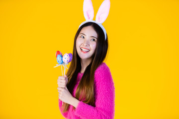 Easter holiday concept, Happy smile Asian Young woman wearing bunny ears hand holding colorful Easter eggs Skewers In isolated on Yellow blank copy space studio background.