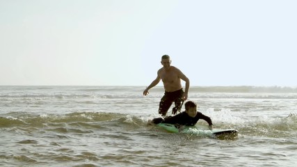 Happy father helping son swimming on board. Cheerful middle aged father helping little son in wetsuit standing and swimming on surfboard on sea waves. Surfing concept