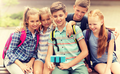 primary education, technology, friendship, childhood and people concept - group of happy school students with backpacks sitting on bench and taking picture by smartphone on selfie stick outdoors