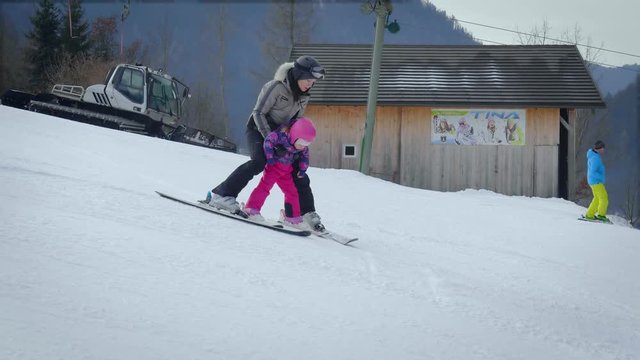 Mother skiing slowly with little girl showing her how to ski downhill on bunny hill