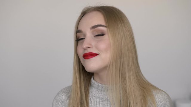 Indoors slow motion close up face shot of young smiling woman with bright red lips, demonstrating trendy makeup by turning face, dressed grey sweater, posing against white wall natural light, model
