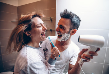 Beautiful young couple in love having fun playing with hair dryer in the bathroom. Husband and wife laughing together at home.