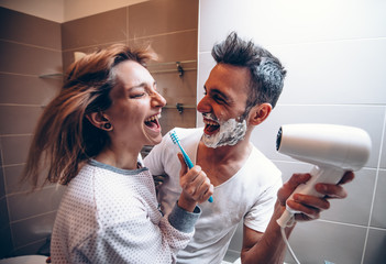 Beautiful young couple in love having fun playing with hair dryer in the bathroom. Husband and wife...