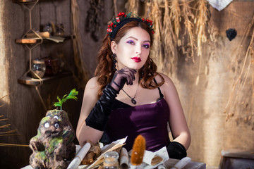 Portrait of a young, attractive witch sitting at a table with magic books and scrolls against a background of alchemical ingredients, flasks, bottles, and dried herbs.