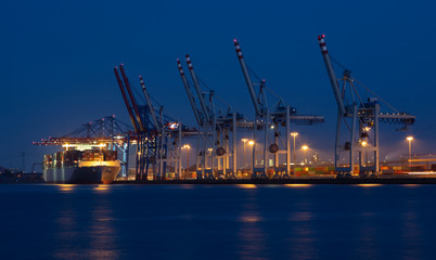 Fototapeta premium A long exposure in the port of Hamburg at night. A container ship lies at the container terminal with many cranes. The lights reflect in the water. Many containers are stacked on land.
