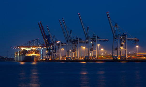 A long exposure in the port of Hamburg at night. A container ship lies at the container terminal with many cranes. The lights reflect in the water. Many containers are stacked on land.