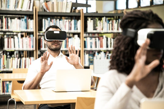 Multiethnic group of students using VR gadgets for studying. Man and woman in virtual reality headsets sitting at desks with laptops and touching air. VR simulators concept
