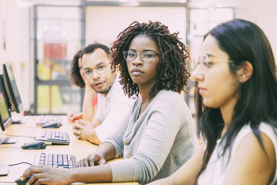 Diverse group of trainees working in computer class. Line of man and women in casual sitting at table, using desktops, looking at monitor or camera. Training concept