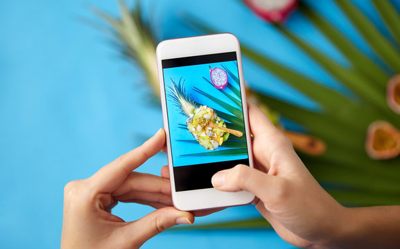 food, technology and healthy eating concept - hands with smartphone taking photo of different exotic fruits mix in bowl with wooden spoon and green fan palm leaf on blue background