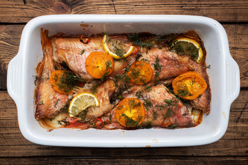 Baked sea bass with tomato pepper lemon and herbs in a ceramic baking sheet on a wooden background....