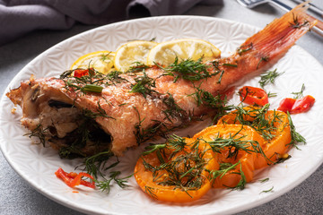 Baked sea bass with tomato pepper lemon and herbs in a ceramic plate on a gray concrete background. Perch fish cooked in the oven.