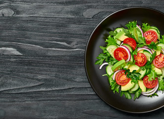 Fresh salad with cherry tomatoes, avocado,cucumeber and lettuce on dish. Healthy vegetarian meal concept. Tasty mixed leaves, tomatoes and mix vegetable salad on black wood, top view.