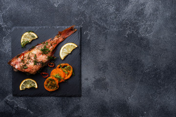 Baked sea bass with tomato pepper lemon and herbs on black slate stand on dark concrete background. Perch fish cooked in the oven. Copy space Top view