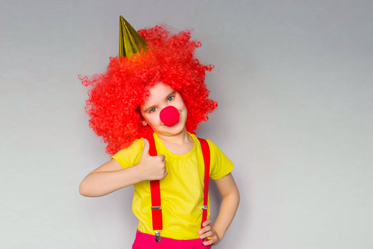 1 April Fool's day concept. Let's celebrate! Funny kid clown playing at home. Girl child with a red wig and a clown's nose is holding a piece of paper for your entry. Copyspace.
