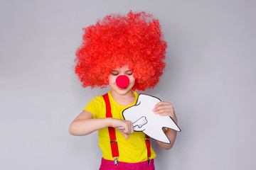 Little funny girl in a red clown wig with a toothbrush and a set of teeth in her hand. Concept of health, oral hygiene, people and beauty. Layout of the room. Selective focus.