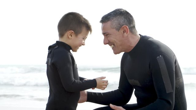 Father and son in wetsuits hugging on beach. Side view of happy father and cite little son talking, laughing and hugging on sea coast, handheld shot. Family time concept