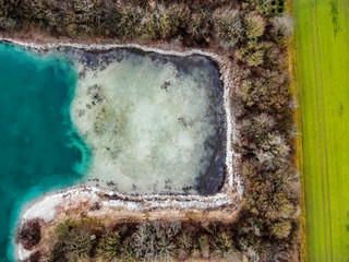 Drohnenbild, Badestrand und Baumreihe an einem türkisen Kiesgrubensee mit kristallklarem Wasser im Winter