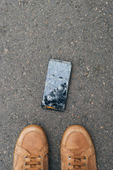 A cell phone dropped on the floor with its owner staring at the phone with its screen broken.