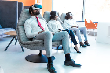 Calm peaceful colleagues watching interactive videos in VR studio. Man and women wearing virtual reality headsets, sitting in motion chairs and relaxing. Visual technology concept